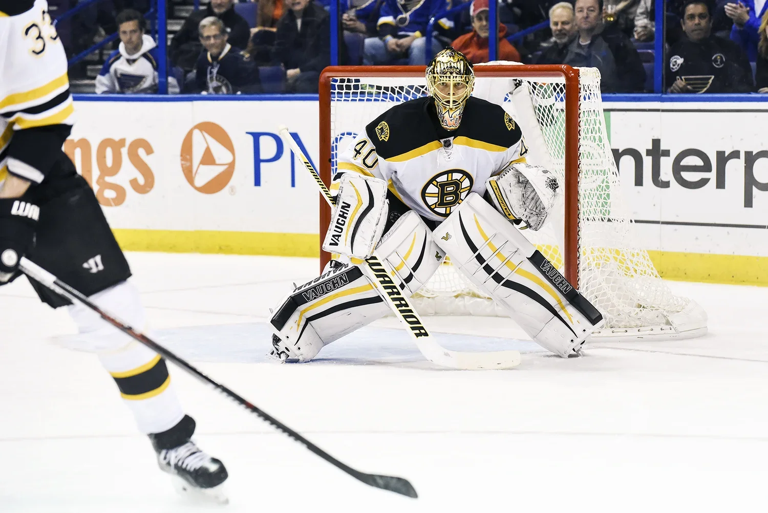 A hockey goalie in a white and black uniform with yellow accents, wearing a helmet with a cage and pads, is standing in front of the goal during a game. Spectators are visible behind the glass.