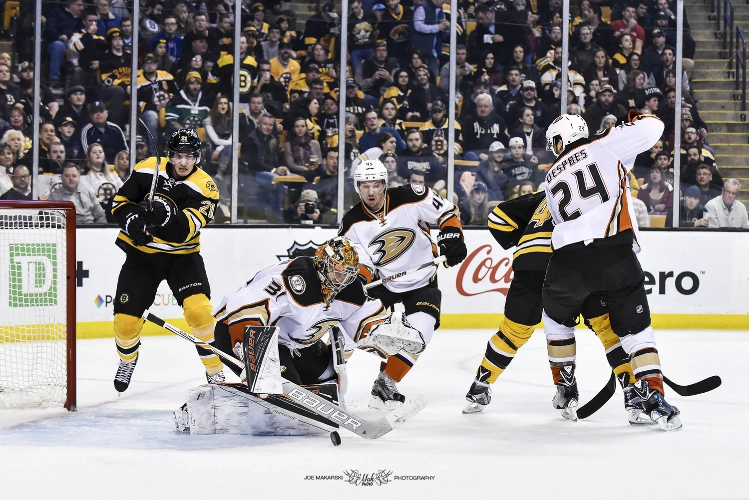 An ice hockey game with players attempting to score a goal. The goalie in white gear is blocking the puck, with players in black and white trying to score. Spectators watch from the stands in the background.
