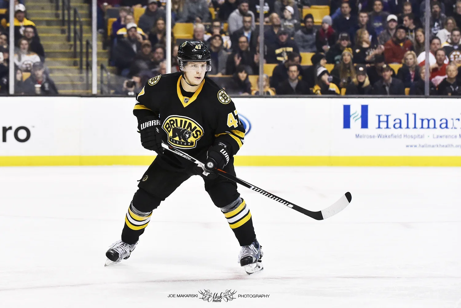 A hockey player in a black Boston Bruins jersey, number 47, is on the ice during a game, with spectators in the stands behind him.