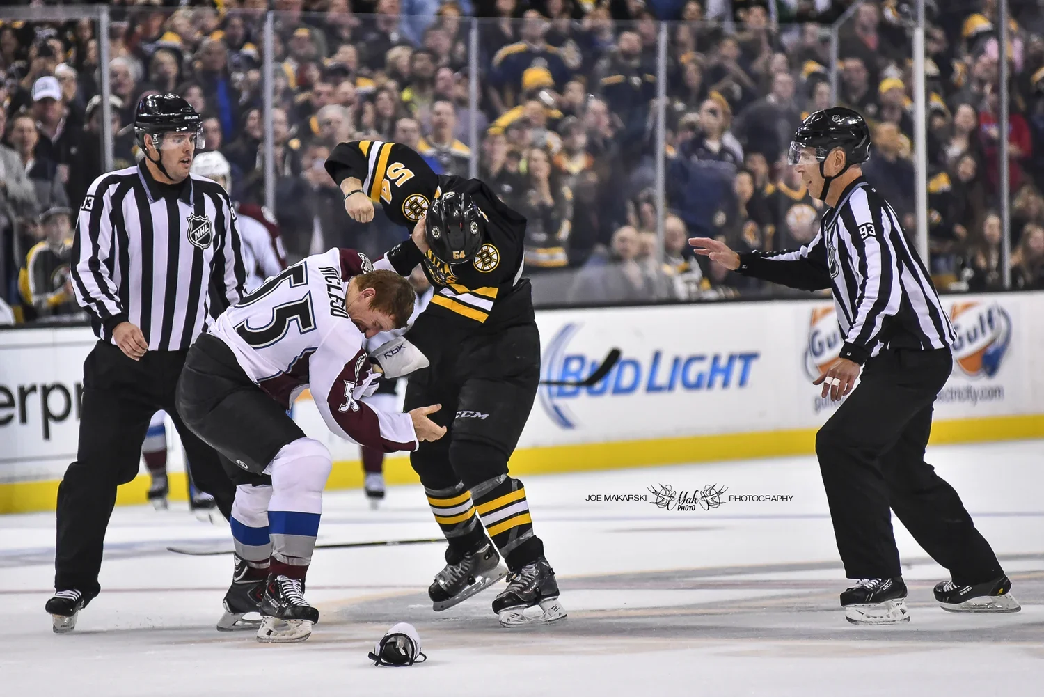 Two hockey players fighting on the ice during a game, with referees trying to intervene and crowd in the background.
