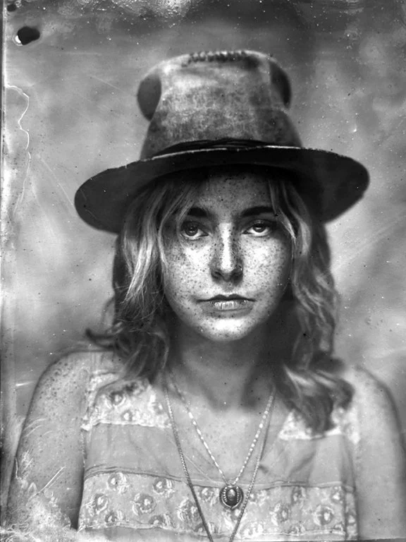 Black and white close-up portrait of a young woman with freckles, wearing a wide-brimmed hat and a patterned top, with layered necklaces.