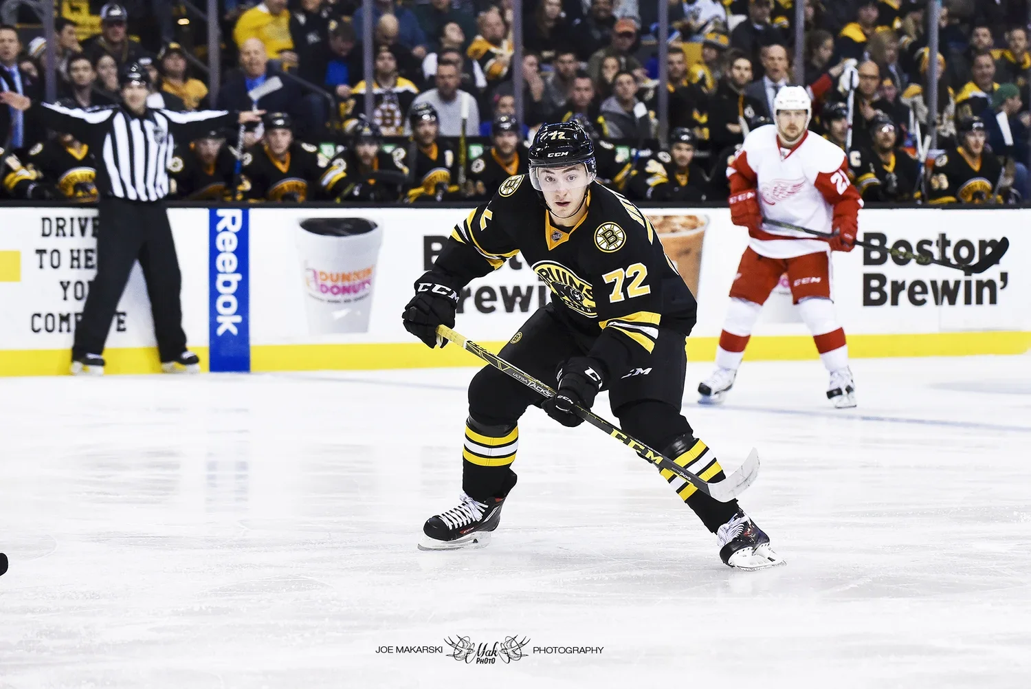 An ice hockey player in a black and yellow jersey with the number 72, skating on the ice with a hockey stick, in front of an audience and a referee, with another player in a white and red jersey in the background.