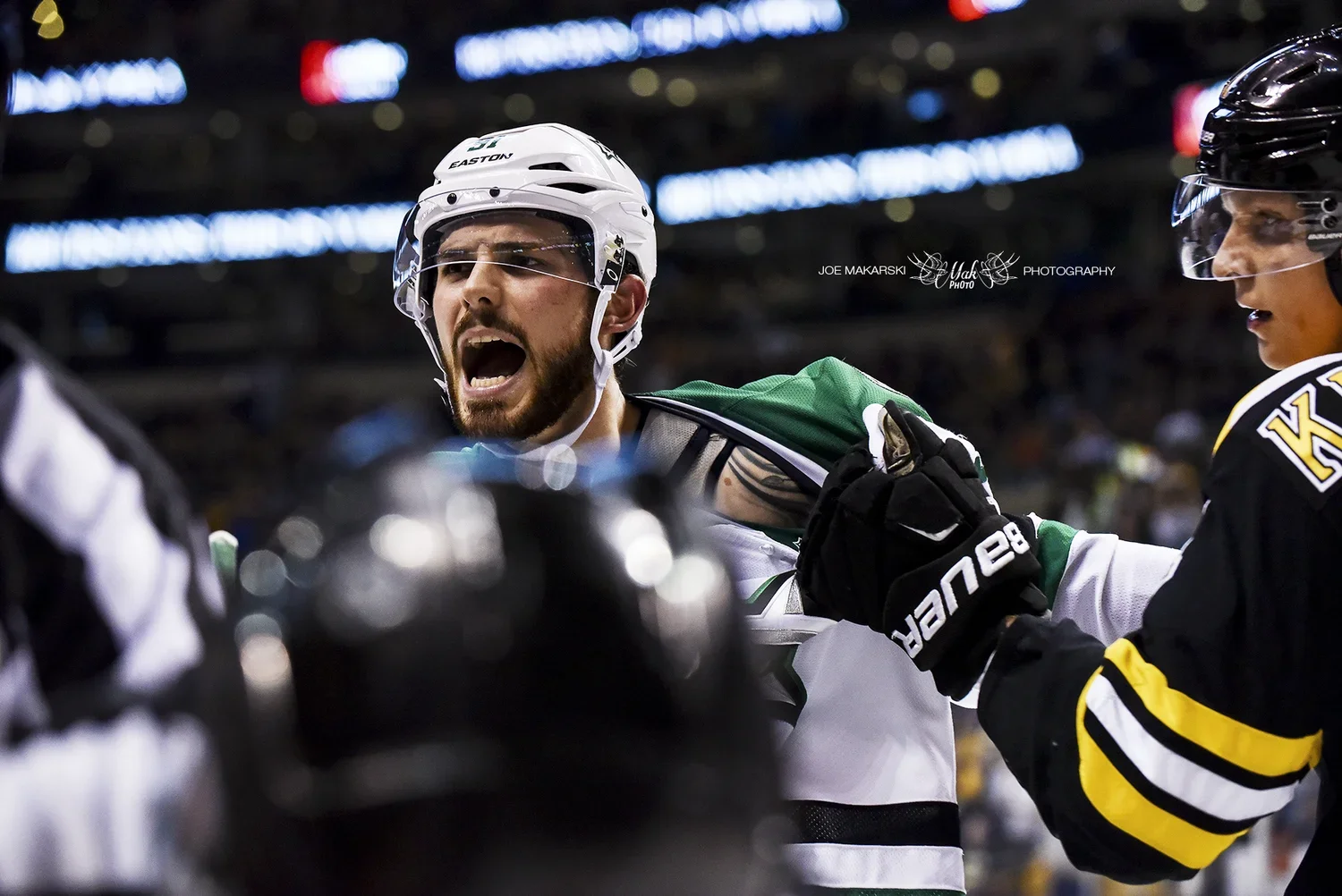 Ice hockey players fighting on the ice rink, one player wearing a green and white jersey and white helmet appears to be shouting, while another player in a black jersey and black helmet is grabbing his jersey.