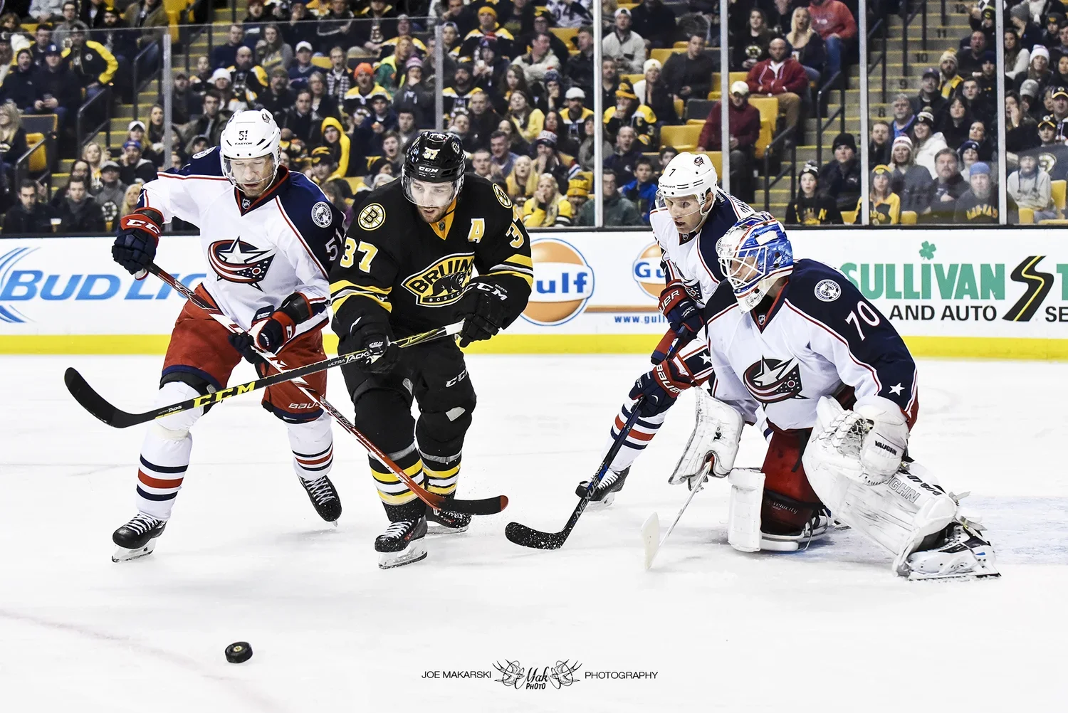 Hockey players in action during a game, with two players from the Columbus Blue Jackets and one from the Boston Bruins try to gain control of the puck near the goal, with a crowd watching in the background.
