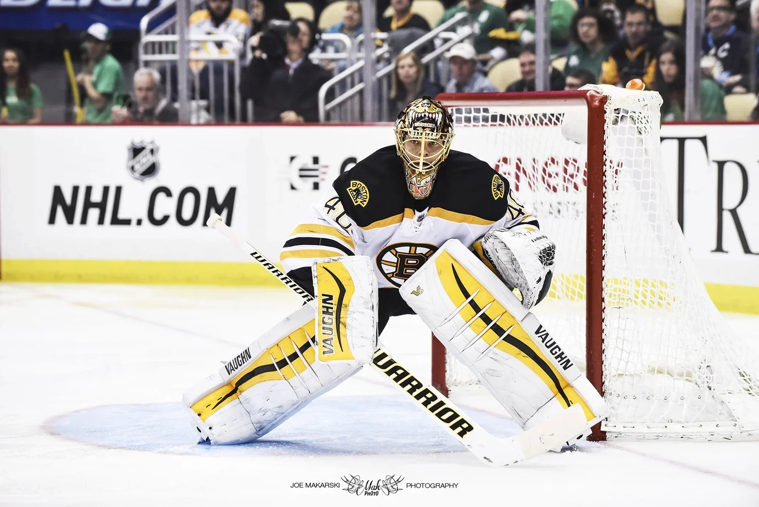An ice hockey goalie in a black and white uniform with yellow accents stands in front of the goal during a game. The goalie wears a mask, goalie pads, and gloves, and is positioned with knees bent and ready to defend the net. Spectators are visible i
