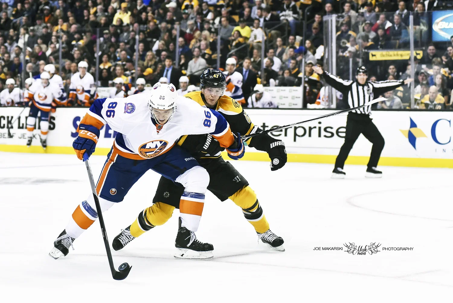 Two ice hockey players, one from the New York Islanders in a white jersey and one from the Boston Bruins in a black and yellow jersey, competing for the puck during a game on the ice rink. A linesman in black and white stripes is in the background al