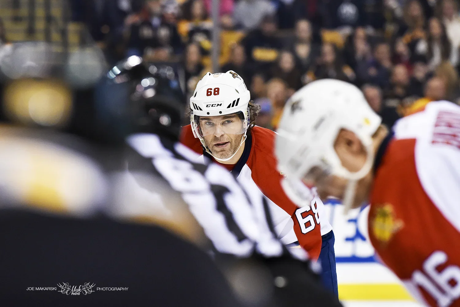 A male ice hockey player wearing a white helmet with number 68 and a red and white jersey, looking intently during a game, with a blurred crowd in the background.