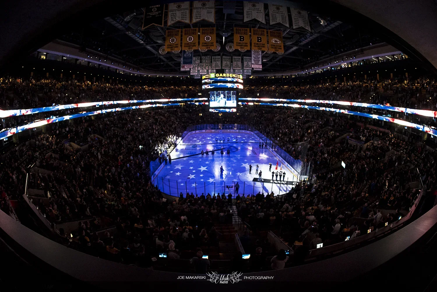 Indoor ice hockey arena filled with spectators, players on the ice, and American flag and stars projection on the ice surface, with banners hanging from the ceiling.