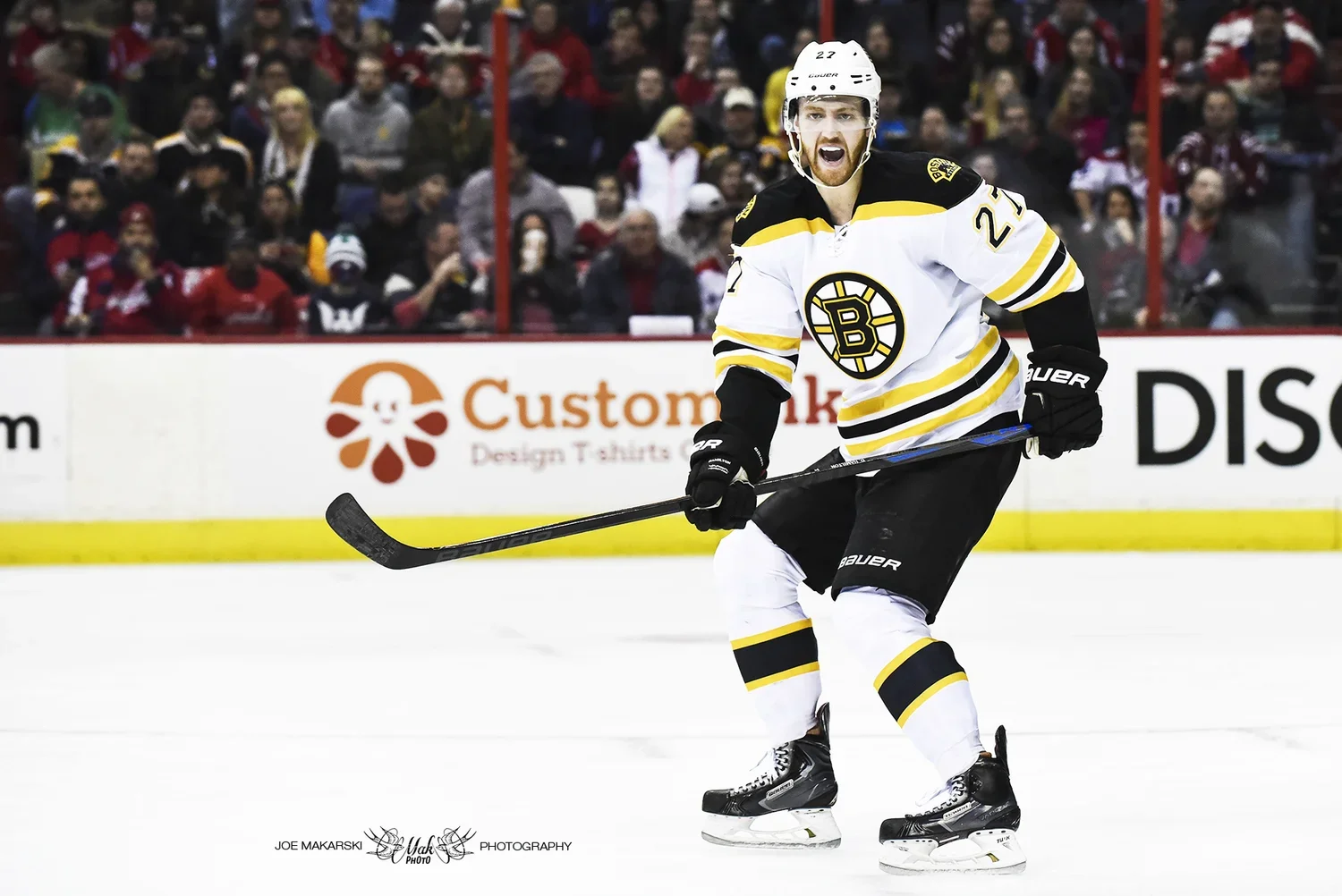 A hockey player in a black, white, and yellow Boston Bruins jersey, holding a hockey stick, skating on the ice during a game with a crowd in the background.