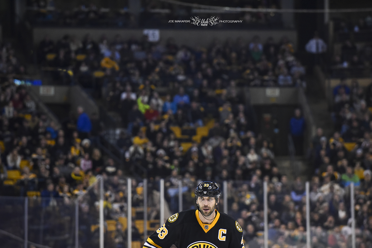 A hockey player wearing a black and yellow jersey and a captain's 'C' on his chest skating on the ice in front of an audience at an indoor hockey arena.