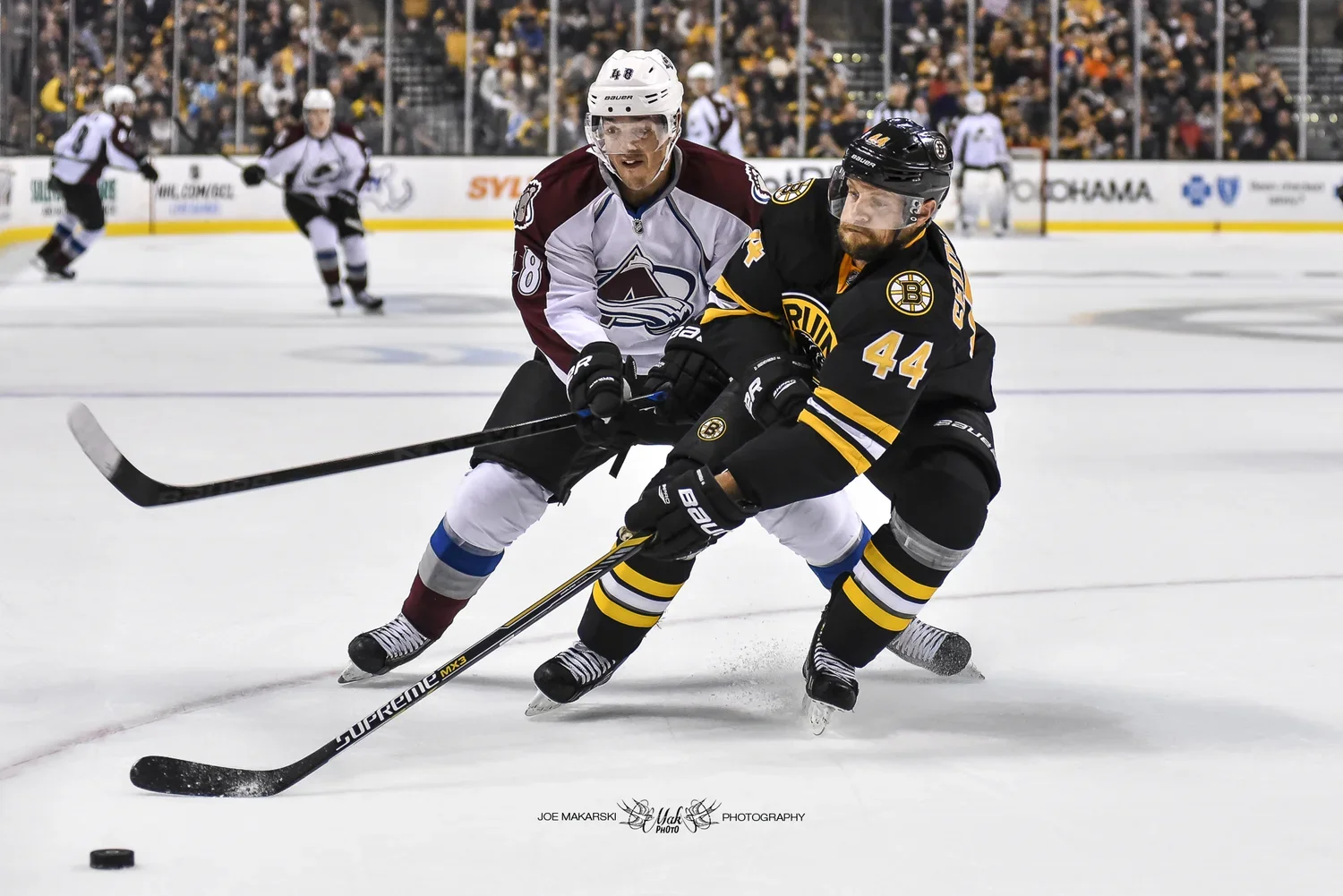 Two ice hockey players in black and maroon uniforms compete for the puck on the ice rink in front of a crowd.
