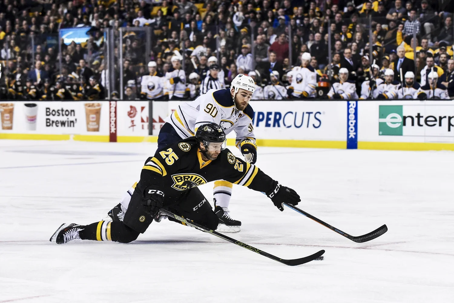 Two ice hockey players competing for the puck during a game, one wearing a black and yellow uniform from the Boston Bruins and the other in a white and blue uniform from the St. Louis Blues, with spectators in the background.