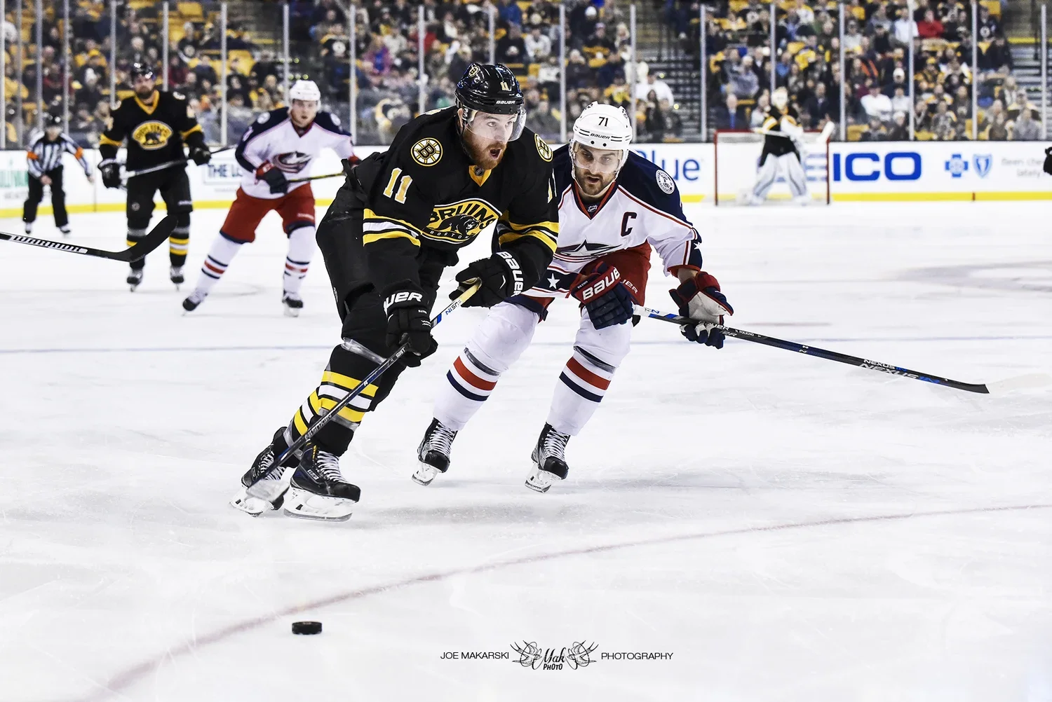 Two hockey players compete for the puck during a game, with additional players and audience in the background.