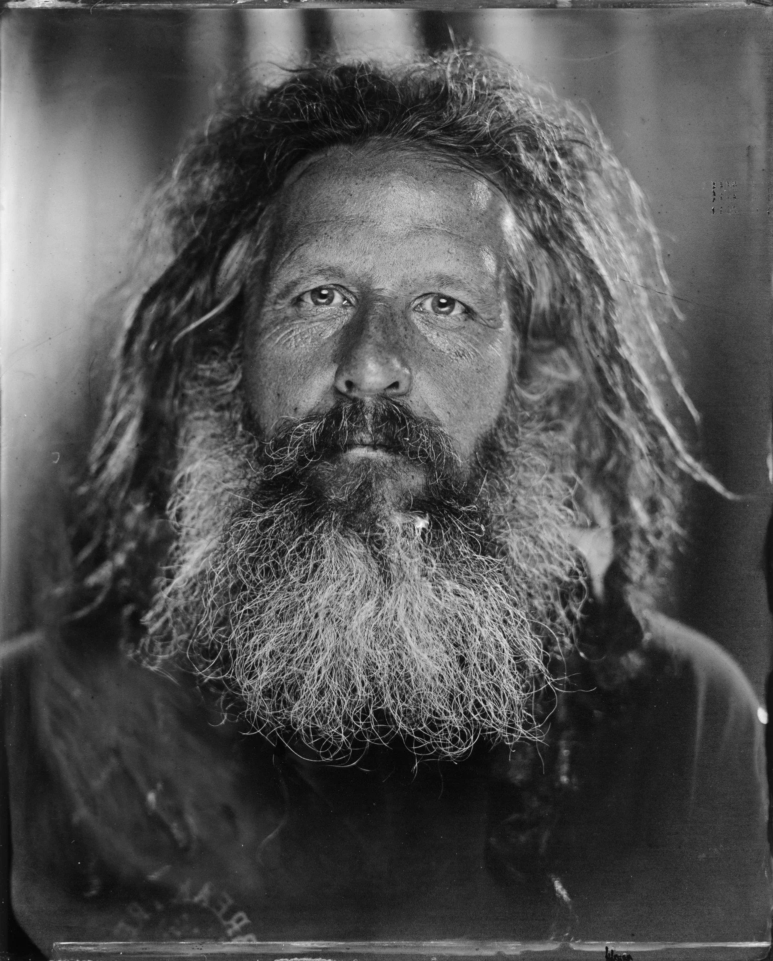 A black and white photograph of an elderly man with long, unkempt hair and a full beard, looking directly at the camera.