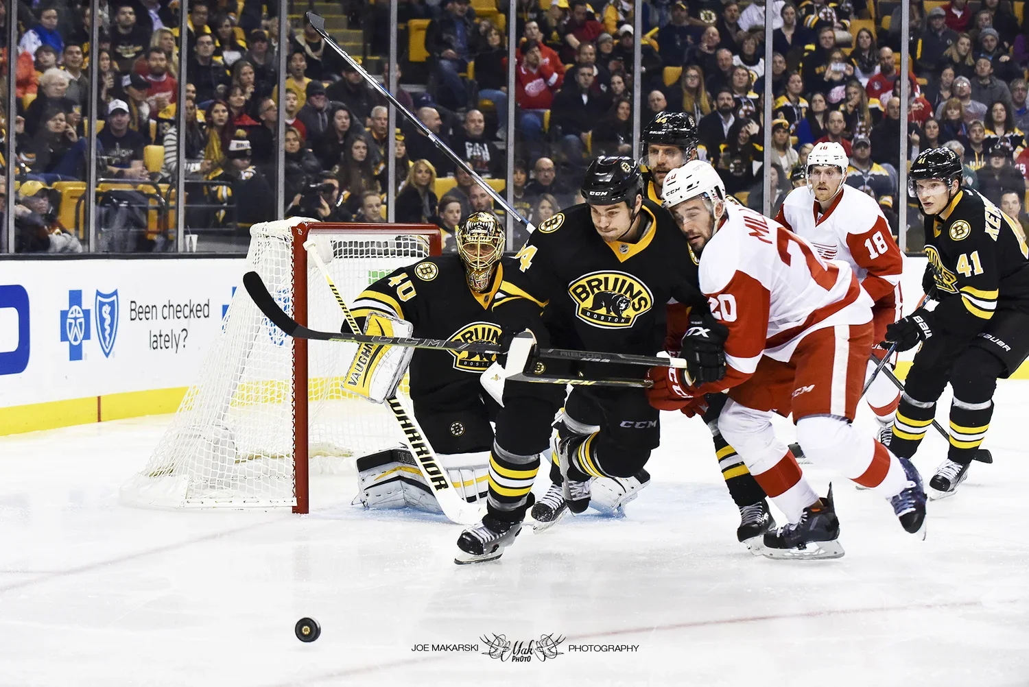 Ice hockey game in progress with players from the Boston Bruins in black jerseys and Detroit Red Wings in white and red jerseys fighting for puck control near the goal, with a crowd watching in the background.