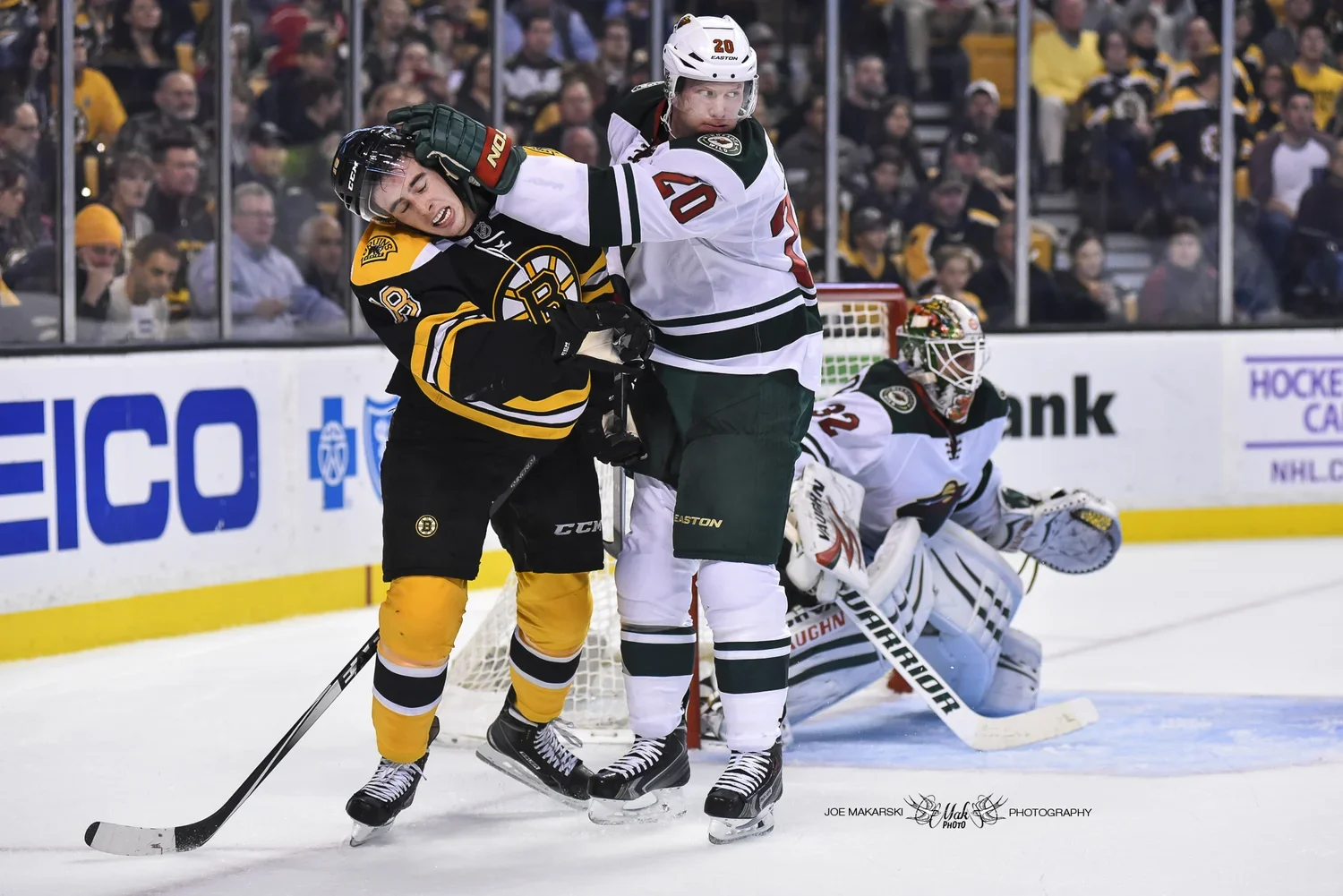 Two ice hockey players, one from the Boston Bruins in black and yellow and the other from the Minnesota Wild in white and green, engage in a fight on the ice, with the Wild player grasping the Bruins player and the Wild goalie in the background prepa