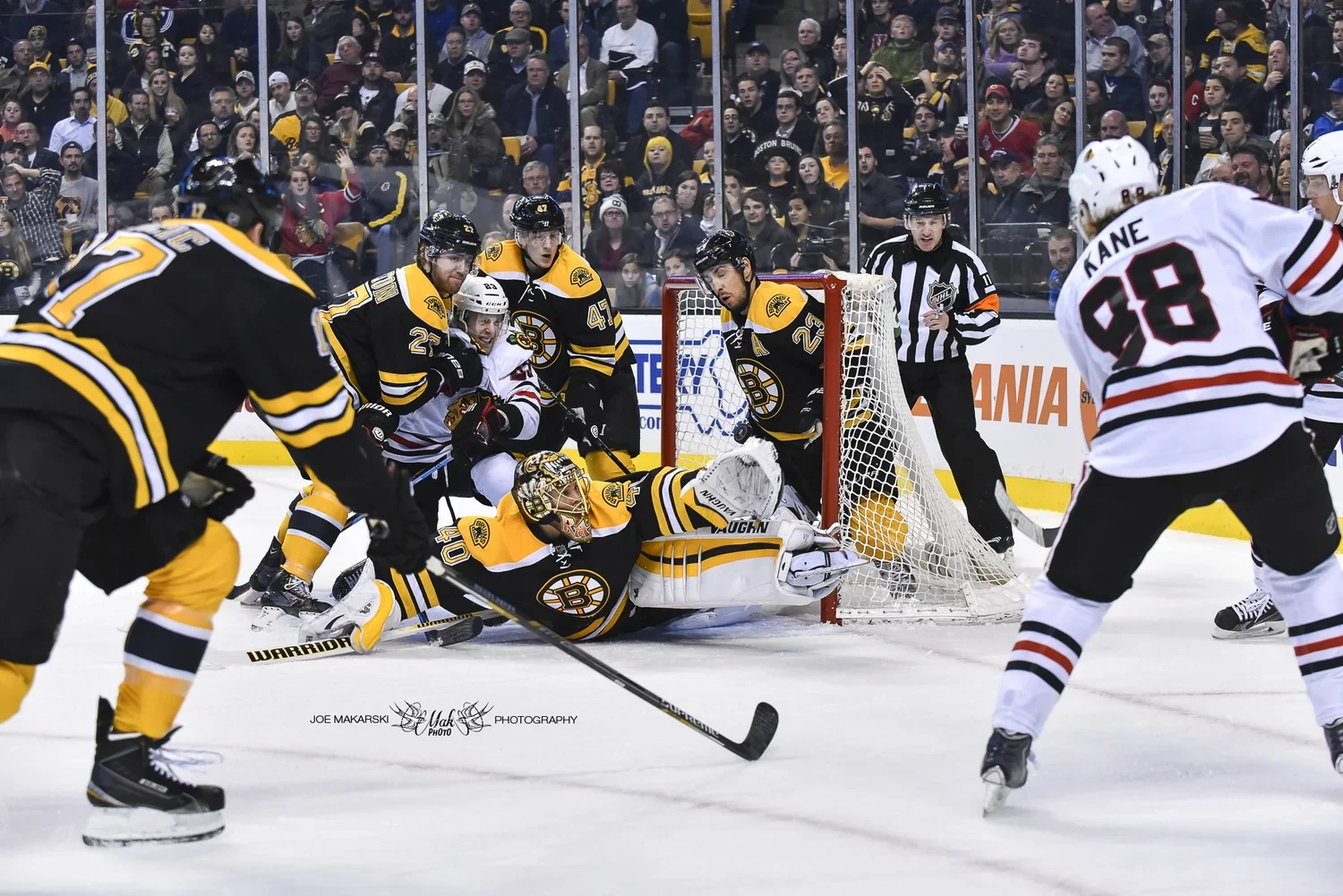 Hockey players in black and yellow uniforms from the Boston Bruins and players in white and red uniforms from the Chicago Blackhawks compete around the goal crease during a hockey game, with the puck near the goalie.