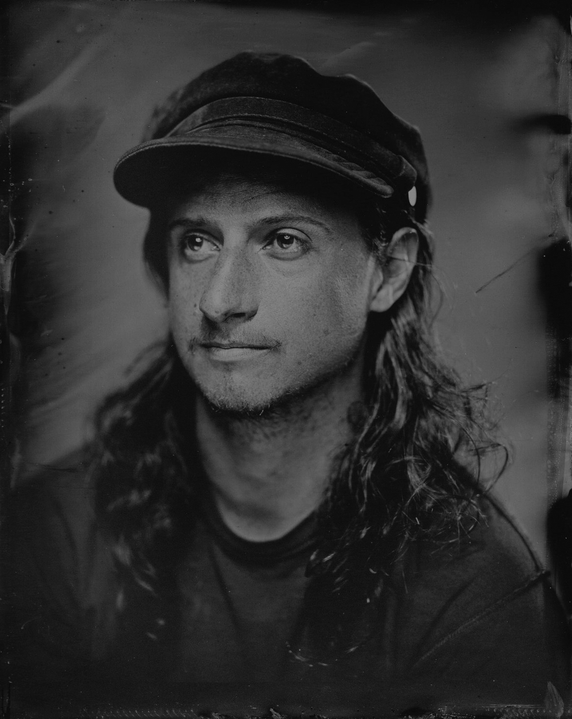 Black and white portrait of a man with long hair, wearing a cap, gazing to the left.