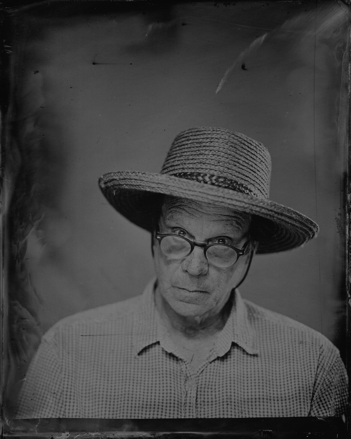 A black-and-white photograph of an elderly man wearing glasses, a checkered shirt, and a wide-brimmed straw hat.