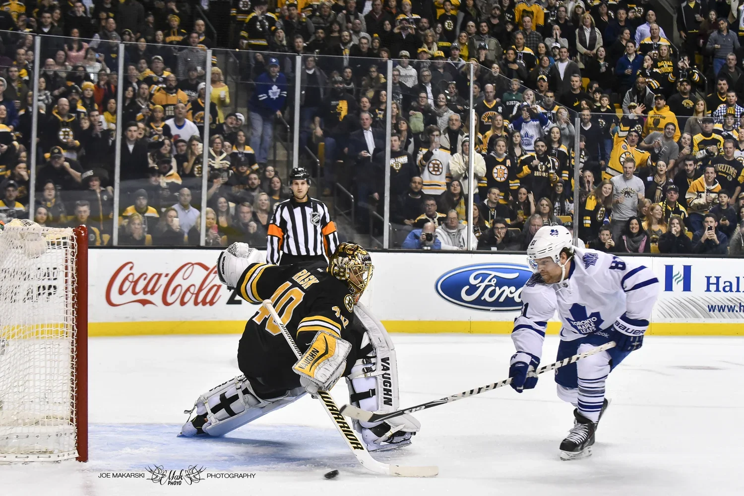 An ice hockey game in progress with a goalie in black and yellow pads near the net, a player in white and blue skating towards the puck, a referee watching, and many spectators in the stands.