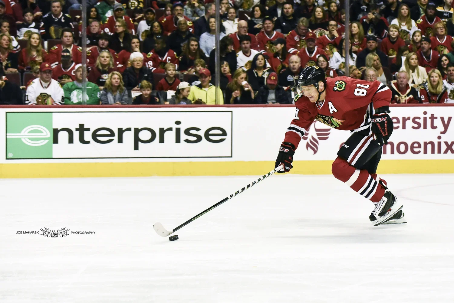 An ice hockey player in a red jersey with the number 18 and an 'A' on their chest, skating on the ice and controlling the puck with their stick, while a crowd of fans in team jerseys watches from the stands.