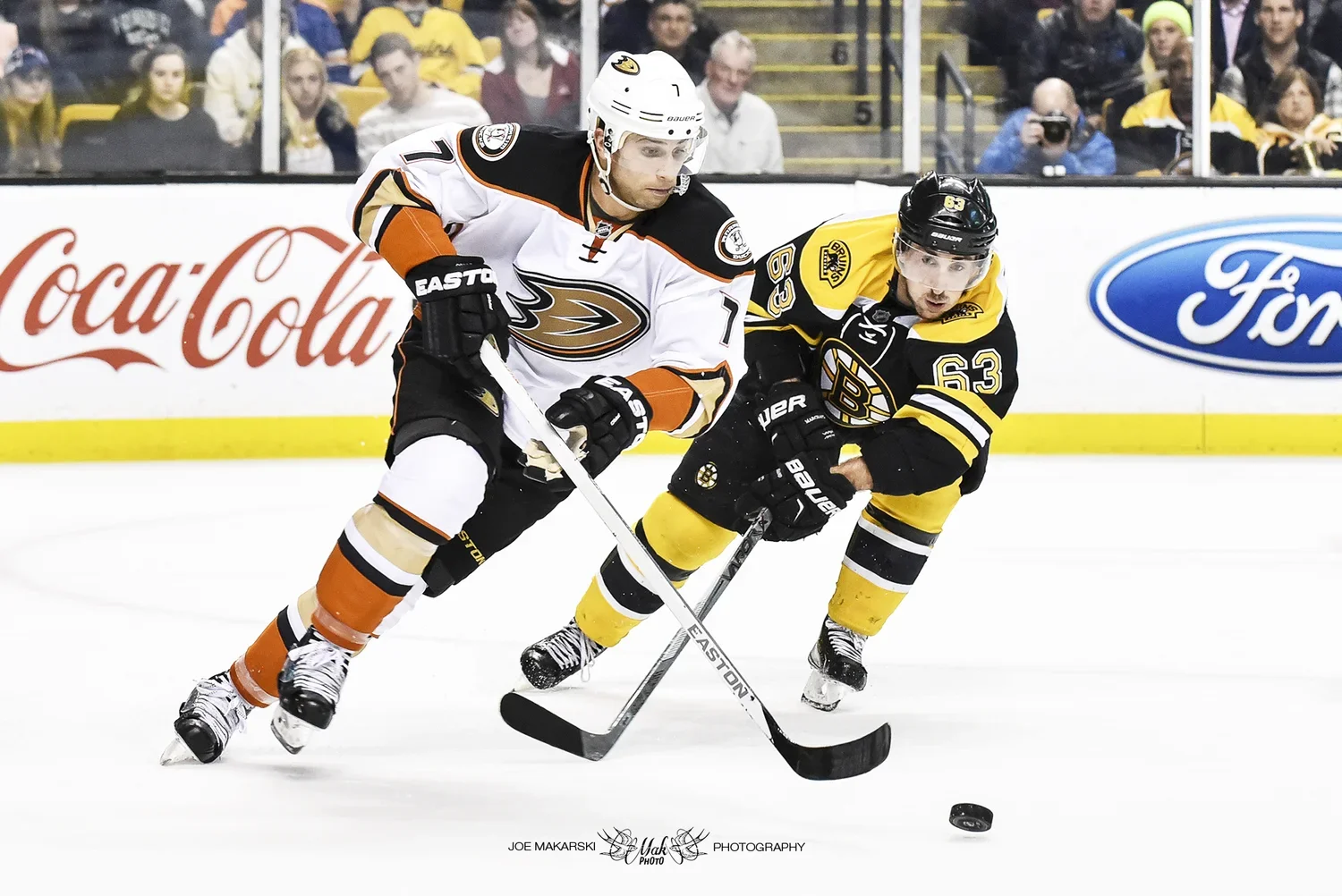 Two hockey players, one in a white and orange uniform and the other in a black and yellow uniform, competing for control of the puck on the ice rink. Spectators watch from the stands in the background.