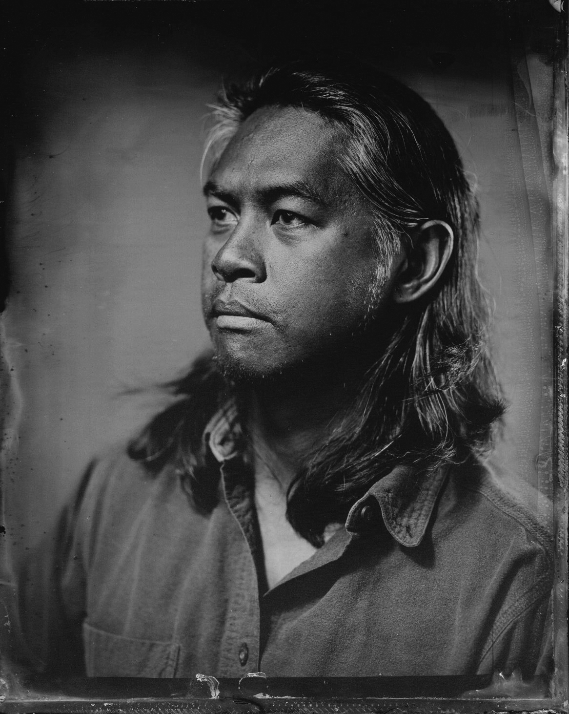 Black and white portrait of a man with long hair, wearing a button-up shirt, looking pensively to the side.