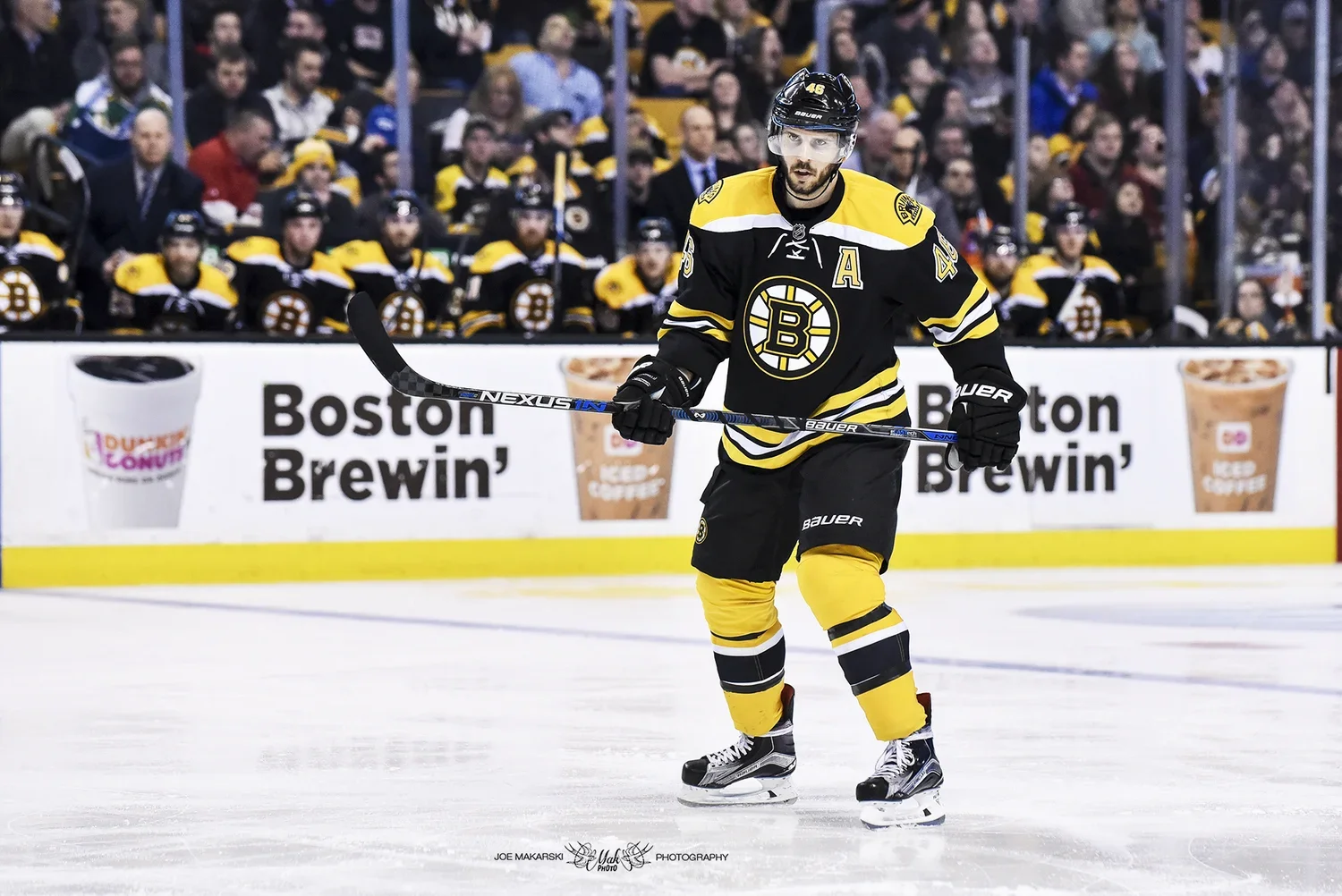 An ice hockey player wearing a black and yellow Boston Bruins uniform skating on the ice during a game, with fans sitting in the background.