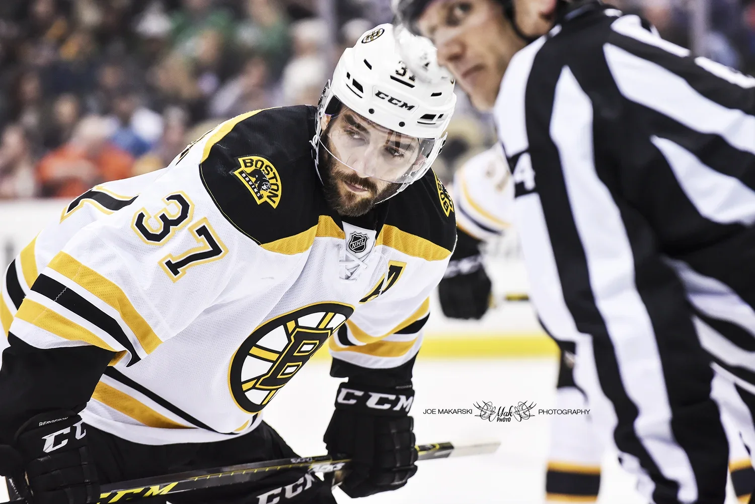 Hockey player in a white and black Boston Bruins jersey with number 37, wearing a helmet and gloves, leaning on the ice and facing a referee.