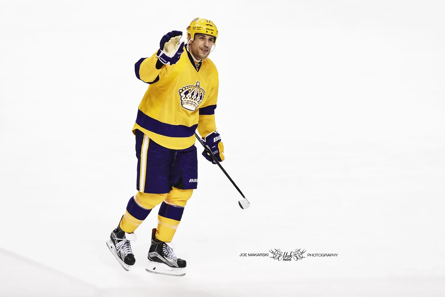 A male ice hockey player in a yellow and purple uniform skating on ice and waving.