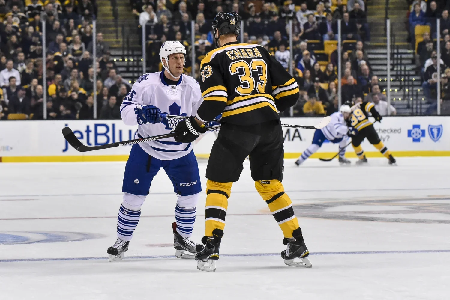 Two ice hockey players, one in a white Toronto Maple Leafs jersey and the other in a black Boston Bruins jersey, face each other on the ice rink during a game. The player in white appears to be holding his stick, while the player in black is standing