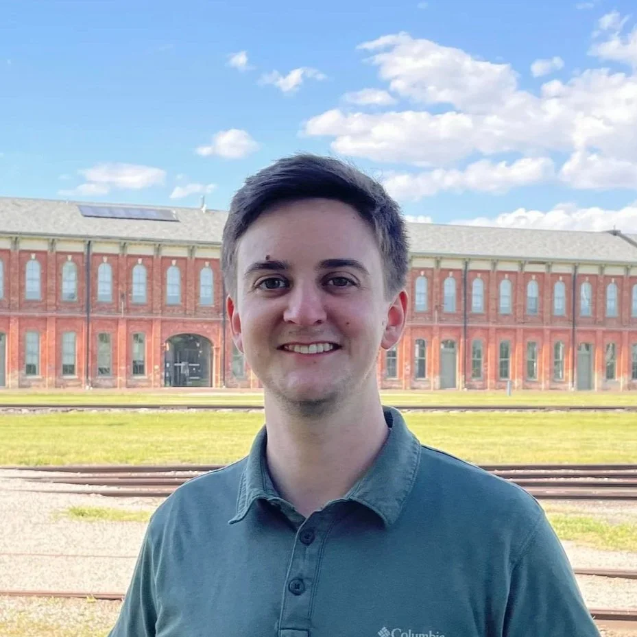 Young man with short dark hair smiling outdoors on a sunny day, with an old brick building and train tracks in the background.