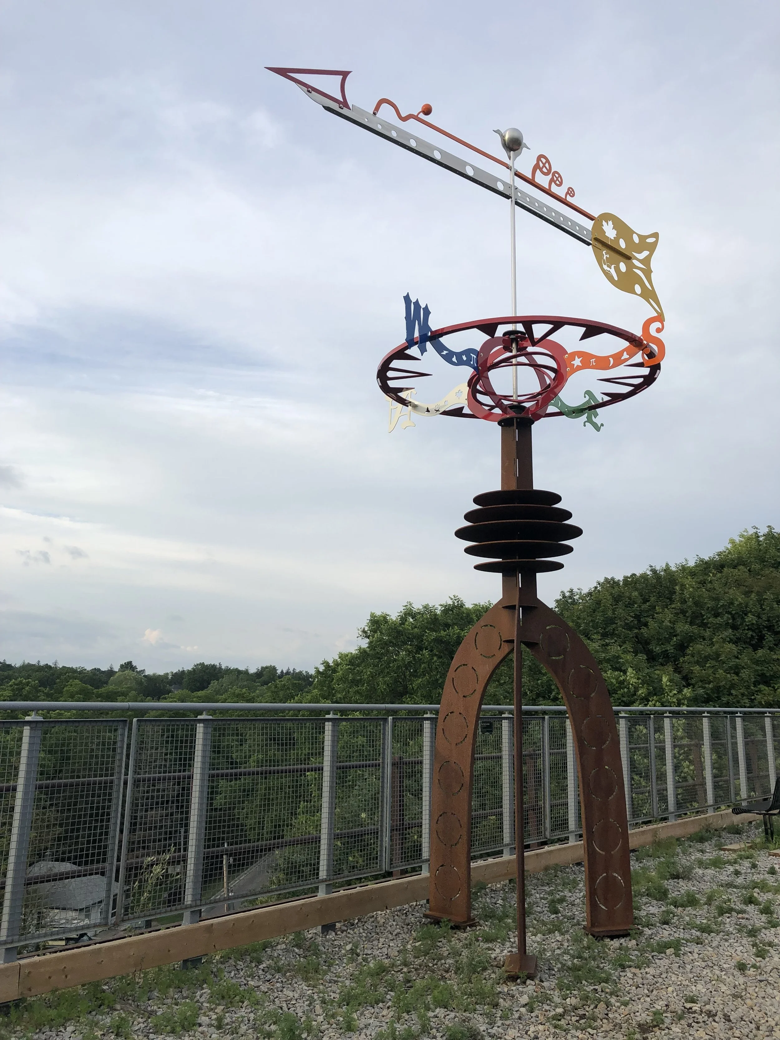 Outdoor sculpture resembling a weather vane with abstract shapes, gears, and leaf patterns, mounted on a tall rust-colored metal stand with a circular support structure. Green trees and a cloudy sky in the background.