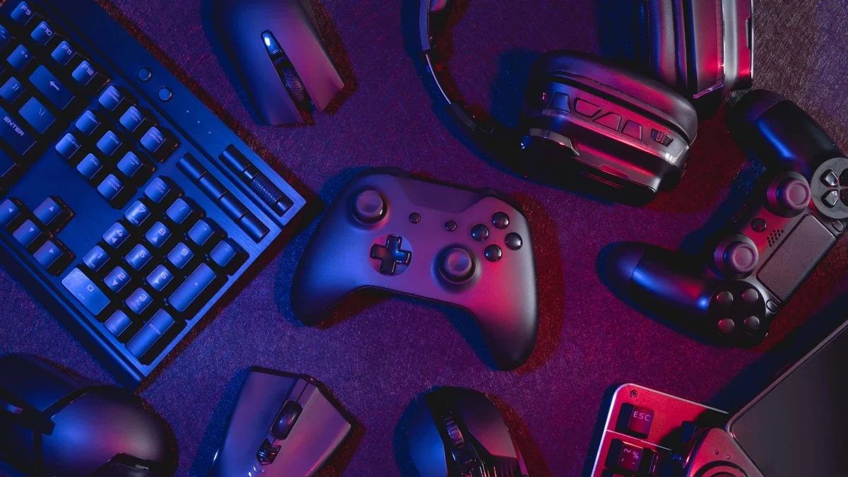 A gaming setup with a black game controller in the center, accompanied by a wireless mouse, a keyboard, two headsets, a PlayStation controller, and a laptop on a dark surface with blue and pink lighting.