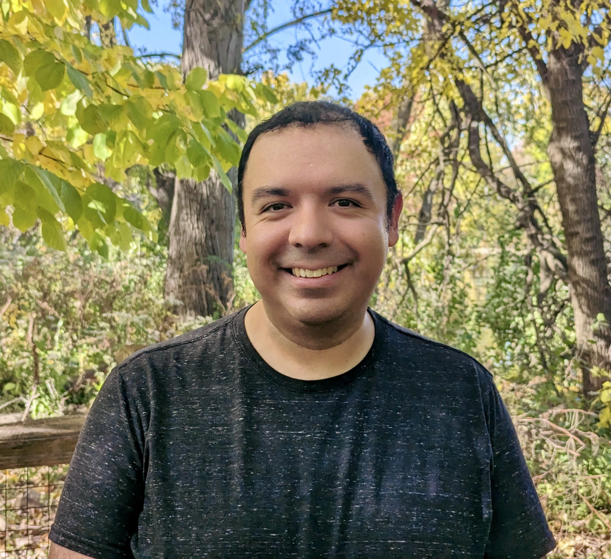 Chris Gonzales, smiling outdoors in a wooded area with green, yellow, and orange foliage on trees
