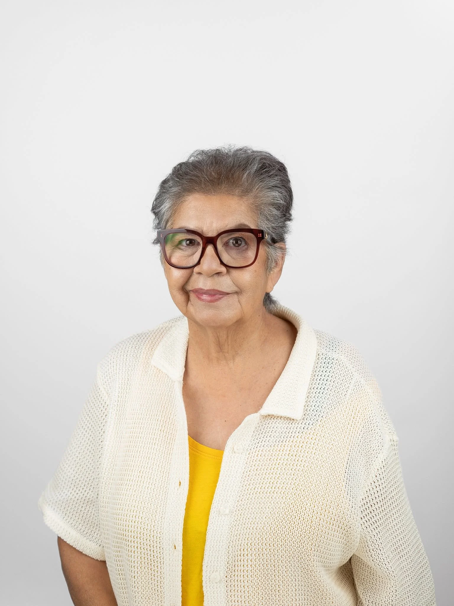 Close-up image of an elderly woman with short gray hair, wearing large glasses, a yellow shirt, and a cream sweater, smiling slightly against a plain white background.