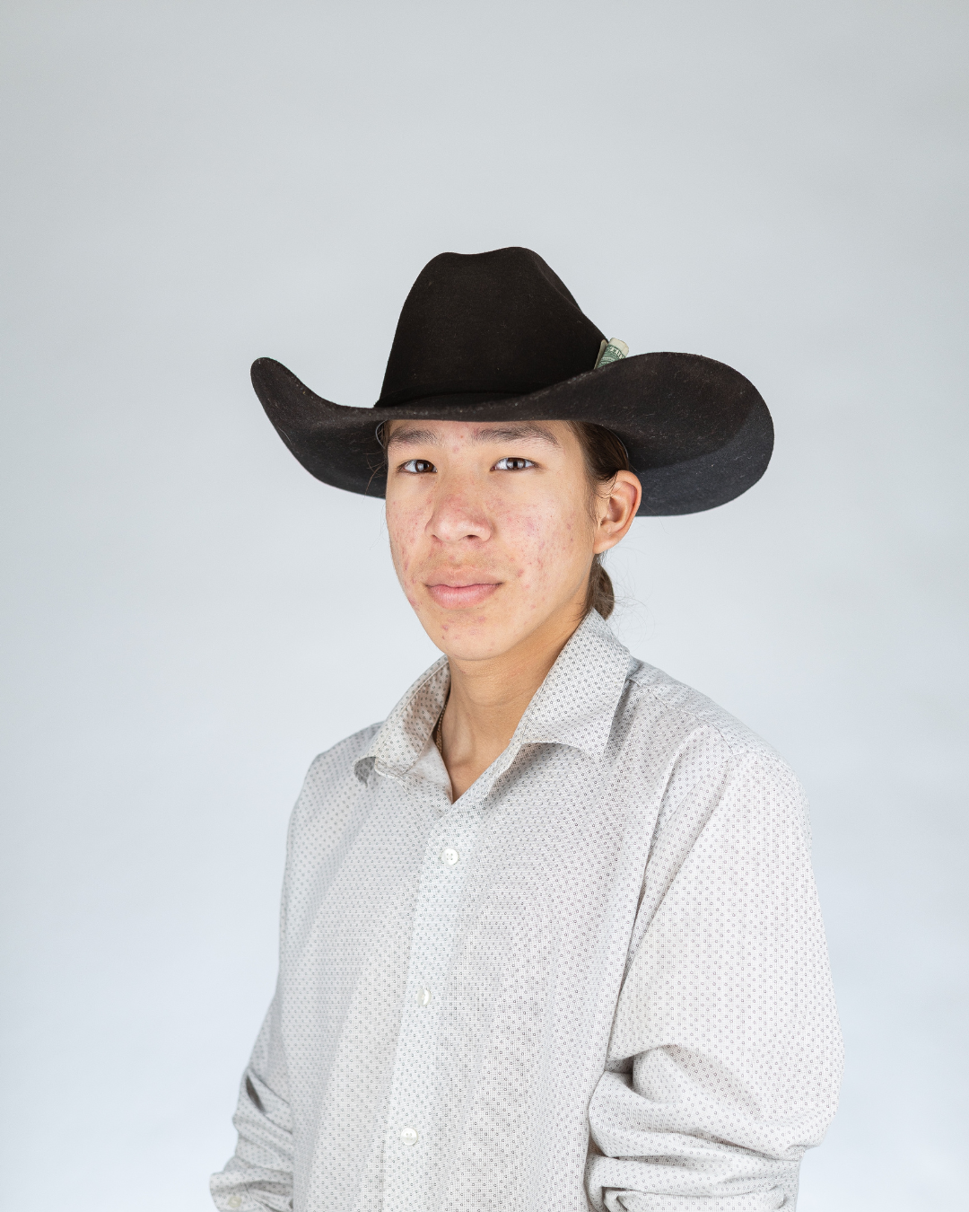 Young person wearing a black cowboy hat and a white button-up shirt with small dots, standing against a plain light background.