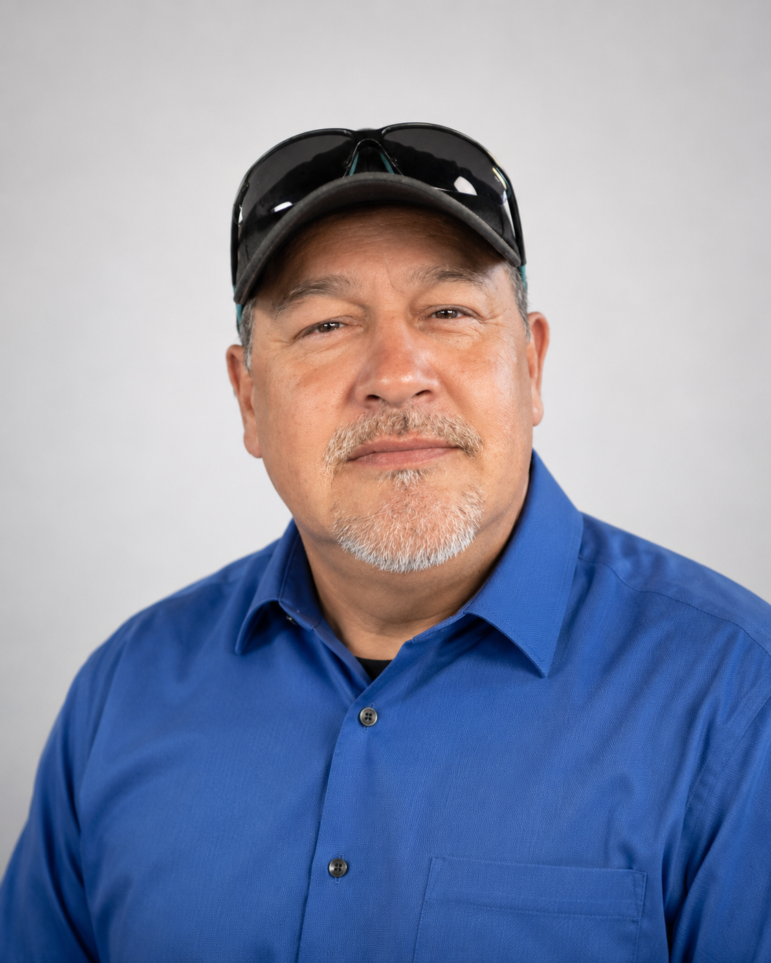 Portrait of a middle-aged man with a goatee, wearing a blue button-down shirt, a black cap, and sunglasses on his head, standing against a plain light background.