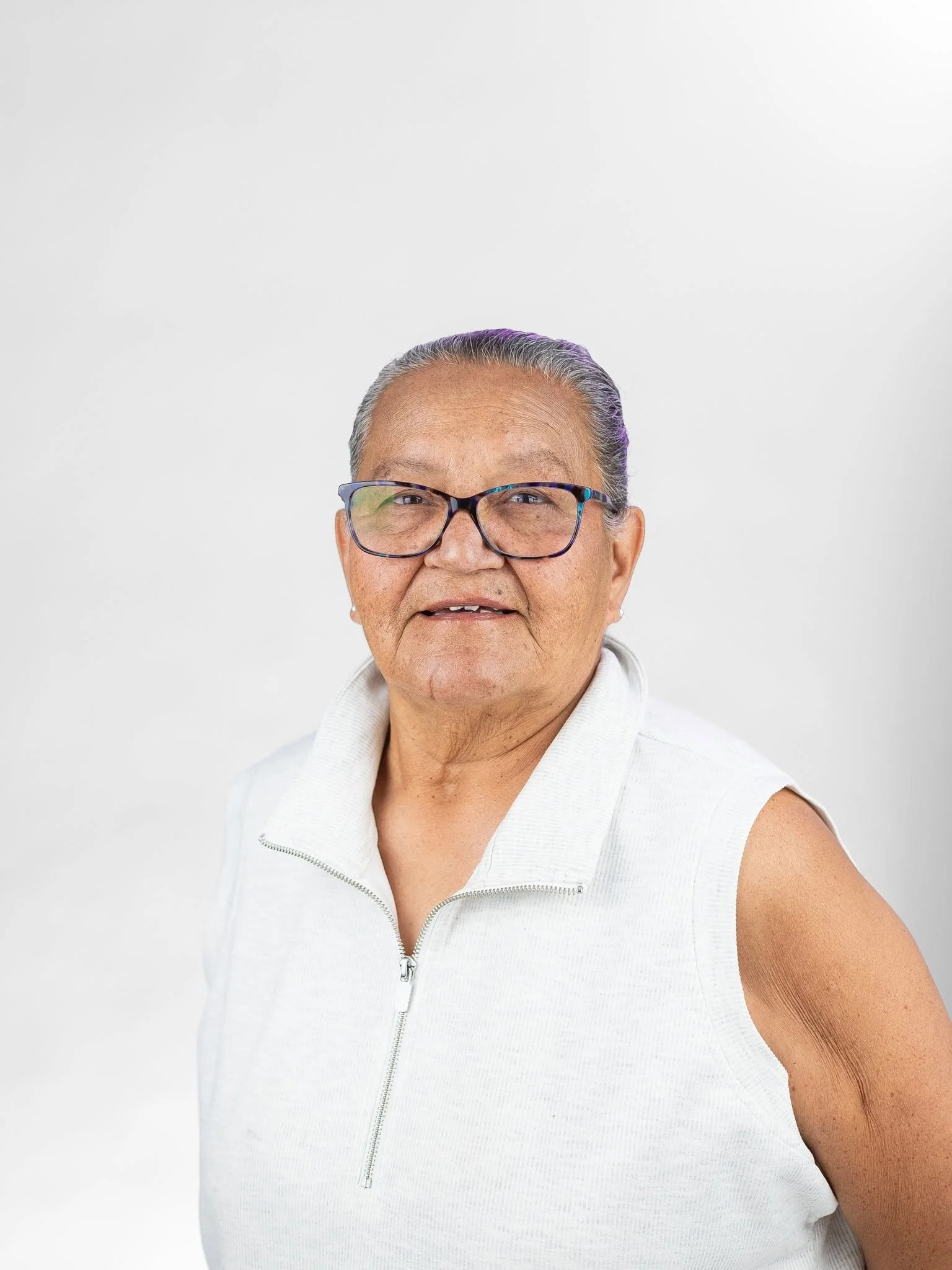 Close-up of an elderly woman with glasses, gray hair styled back, wearing a sleeveless white zip-up vest, smiling against a plain white background.
