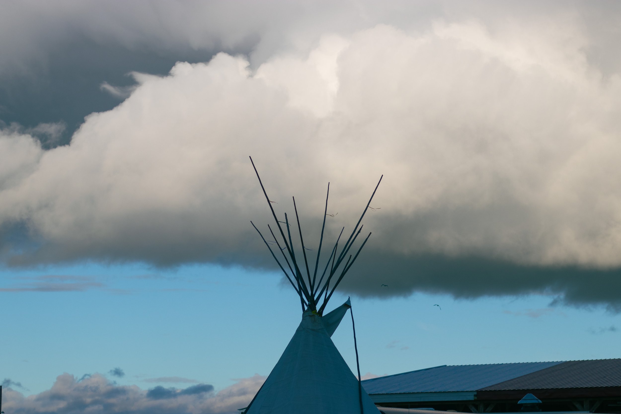 A teepee tent with a sheer fabric cover at the bottom and sticks protruding from the top, set against a cloudy sky with some visible buildings and rooftops in the foreground.