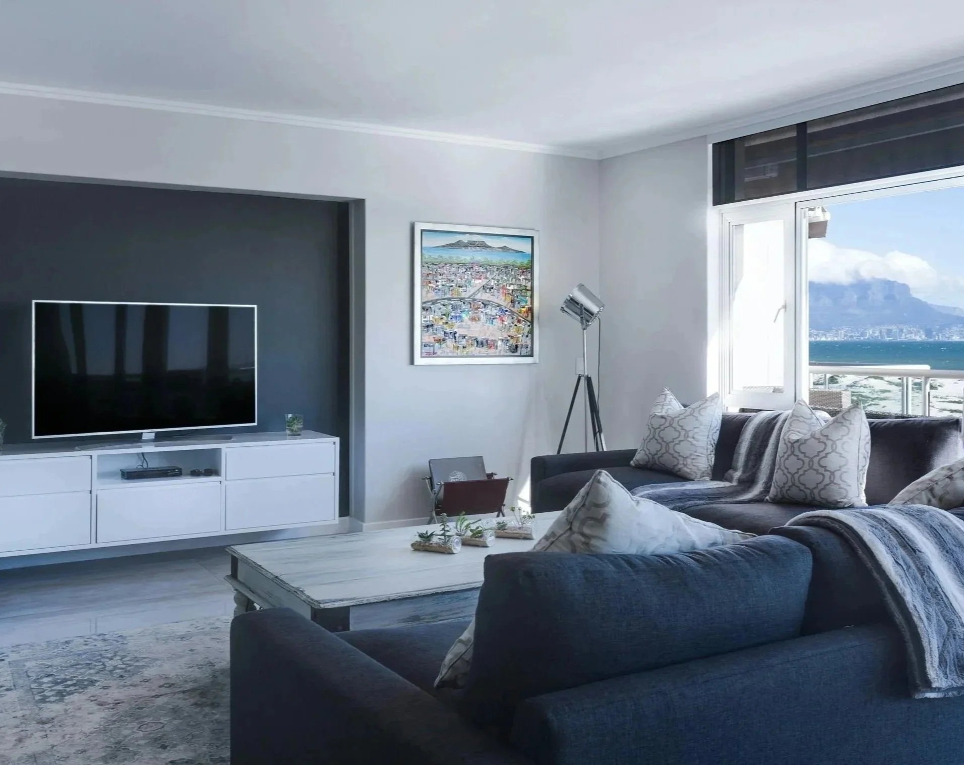 Open accessible living room with dark gray sofa, white coffee table, white media console with a TV, wall art  large window showing a view of the ocean and mountains, floor lamp, and a view of the ocean outside.