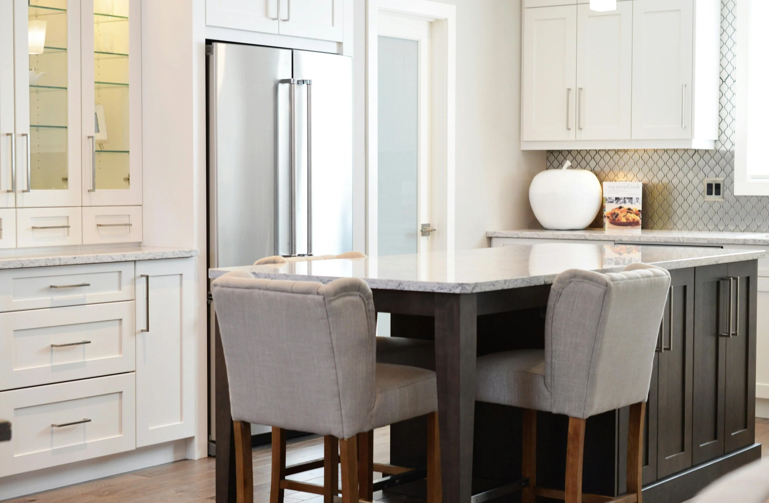 A modern kitchen with white cabinets, a stainless steel refrigerator, a marble countertop island, and four upholstered chairs around the island. There is a decorative vase and a cookbook on the countertop, and the background features a textured backsplash and a frosted glass door.