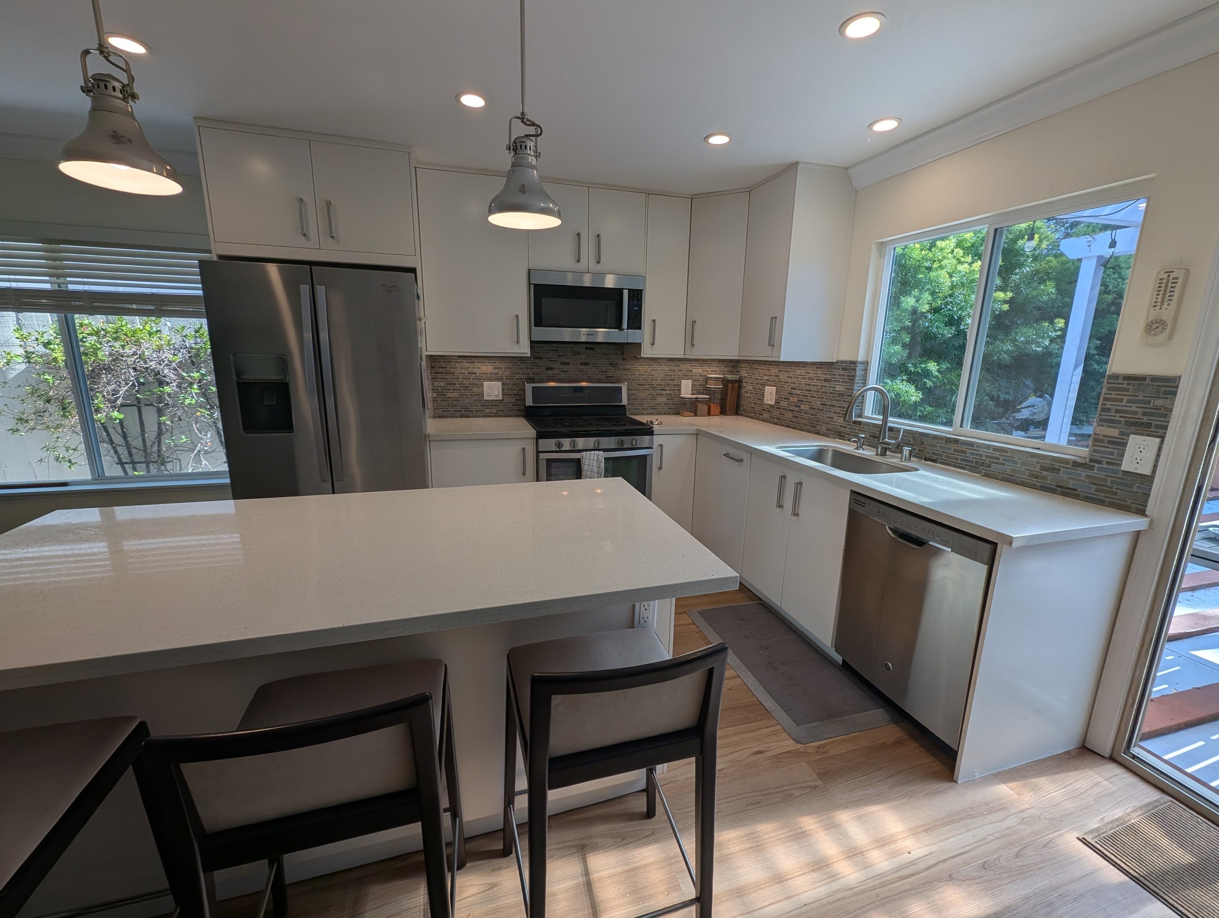 Modern kitchen with white cabinets, stainless steel refrigerator, oven, and microwave, large window with greenery outside, kitchen island with black chairs, hardwood floor, pendant lights, and a sliding glass door.