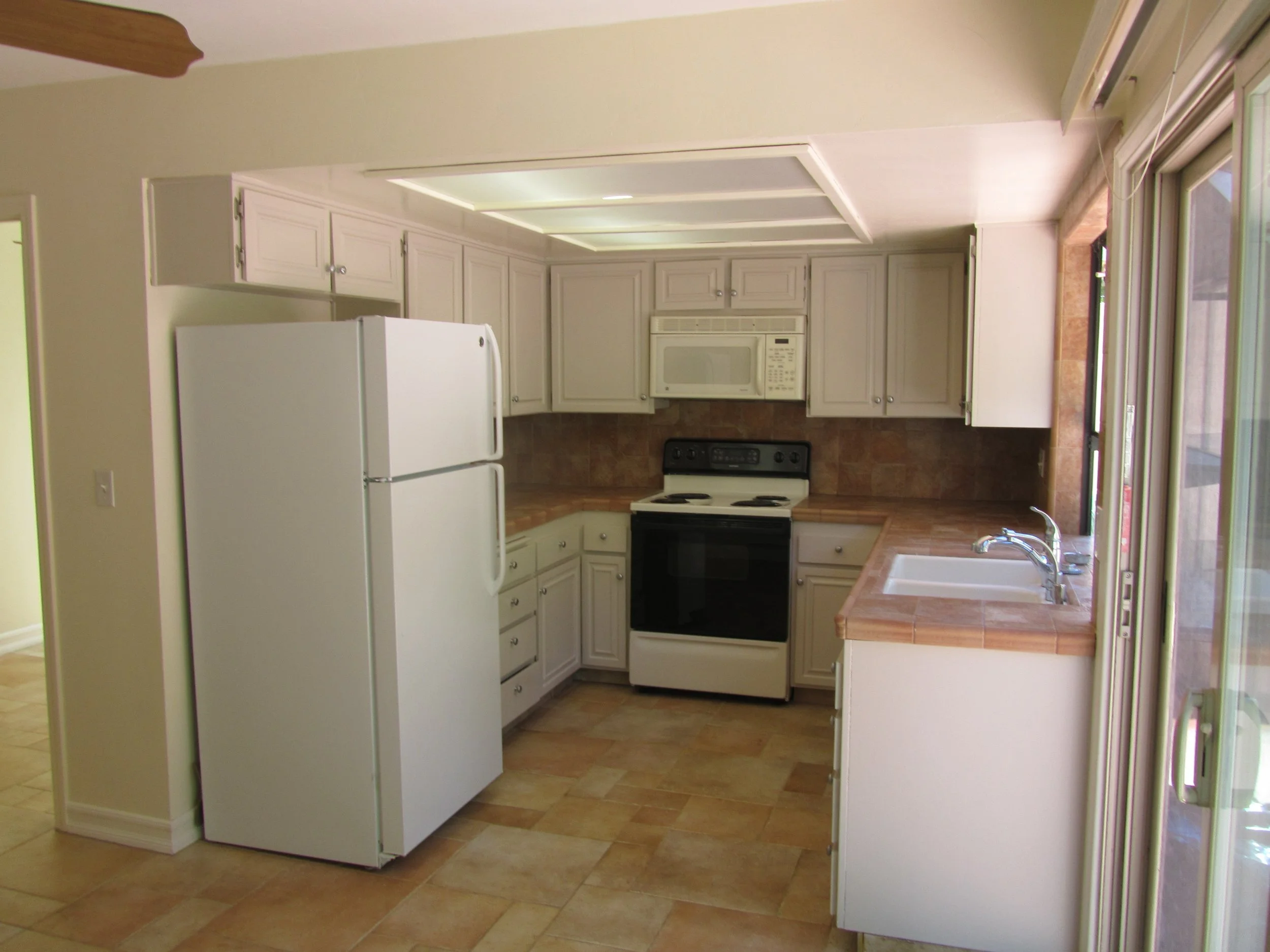 Existing 8 1/2' x 8 1/2' 1970's builder grade kitchen A kitchen with dropped ceiling, recessed fluorescent lighting, white cabinets, a white refrigerator, a white microwave, a black oven, and a double sink under large windows, with tiled flooring.
