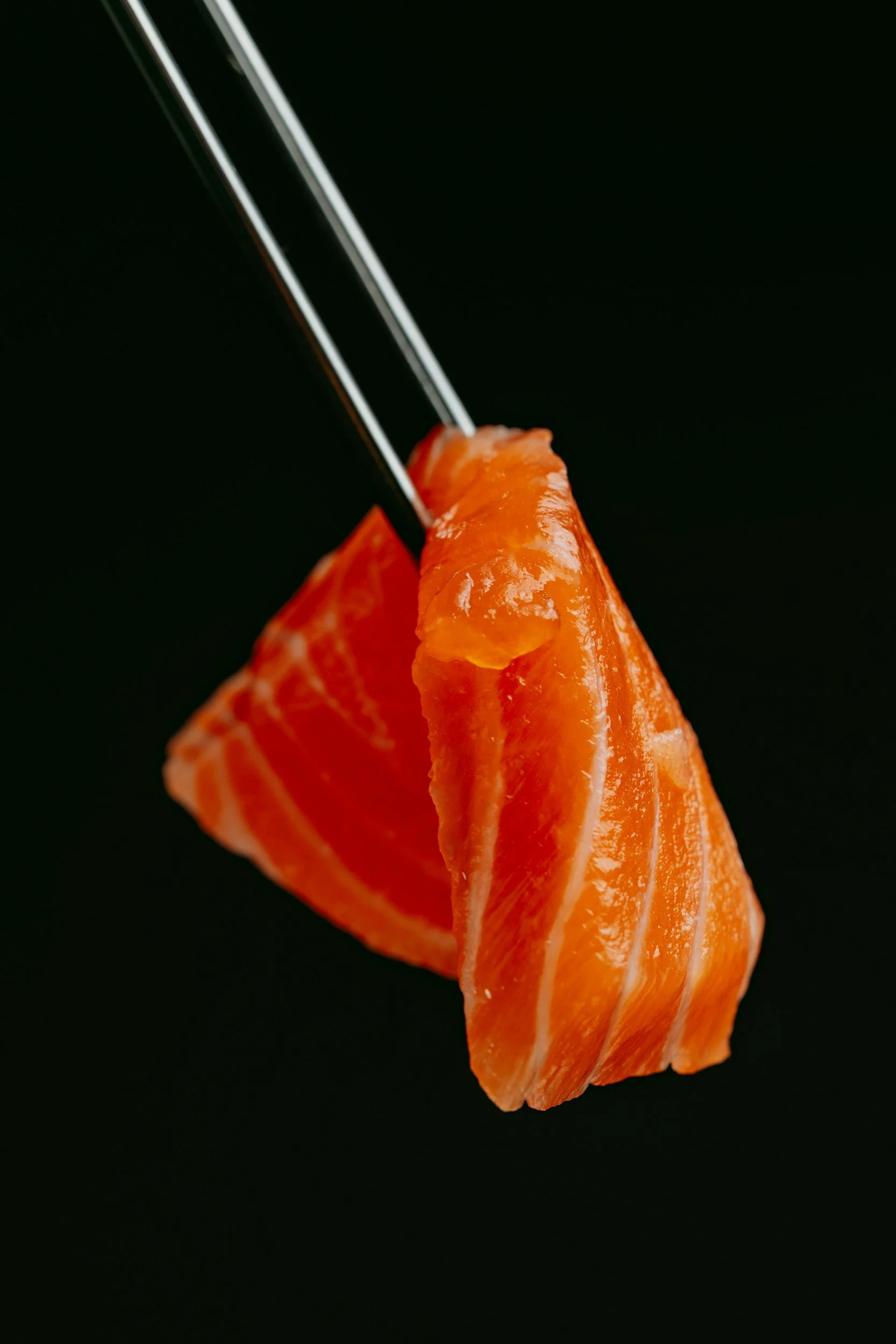 Close-up of a piece of fresh salmon sashimi being held by chopsticks against a black background.