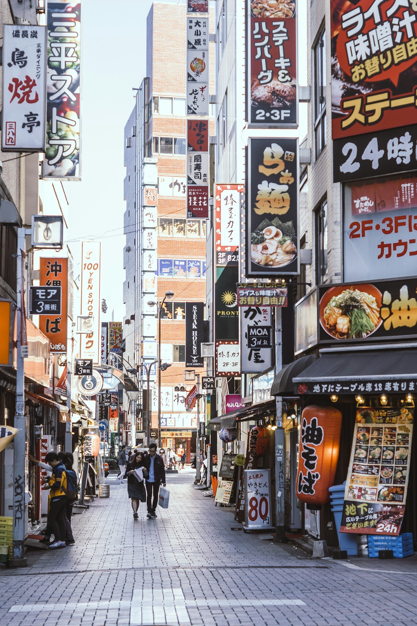 A busy pedestrian street in Japan with numerous signs and storefronts, some advertising food like ramen and seafood, and people walking and shopping.