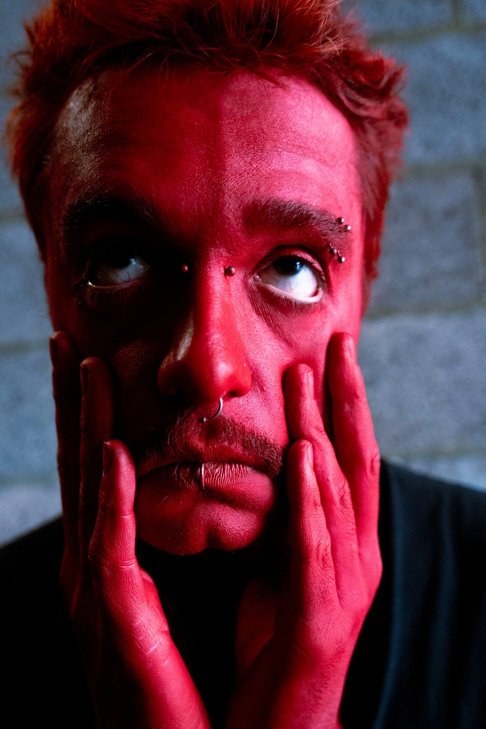 A person with red-painted face and multiple facial piercings, touching their face with their fingers, with a dark background.