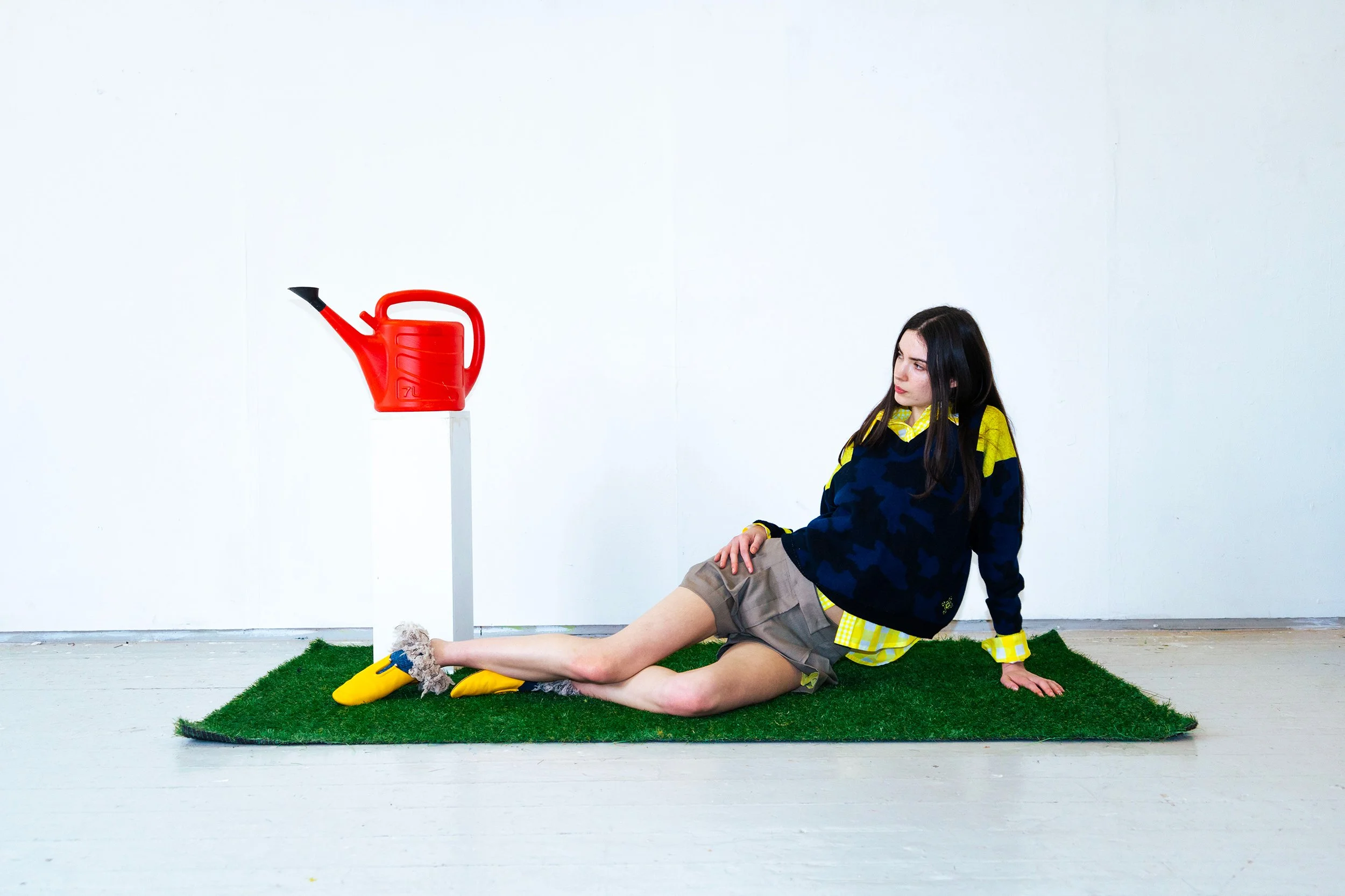 A young woman with dark hair, wearing a yellow and black jacket, gray shorts, and yellow shoes, sitting on an artificial grass mat, leaning back on her hands, with a red watering can on a stand placed nearby, against a white wall.