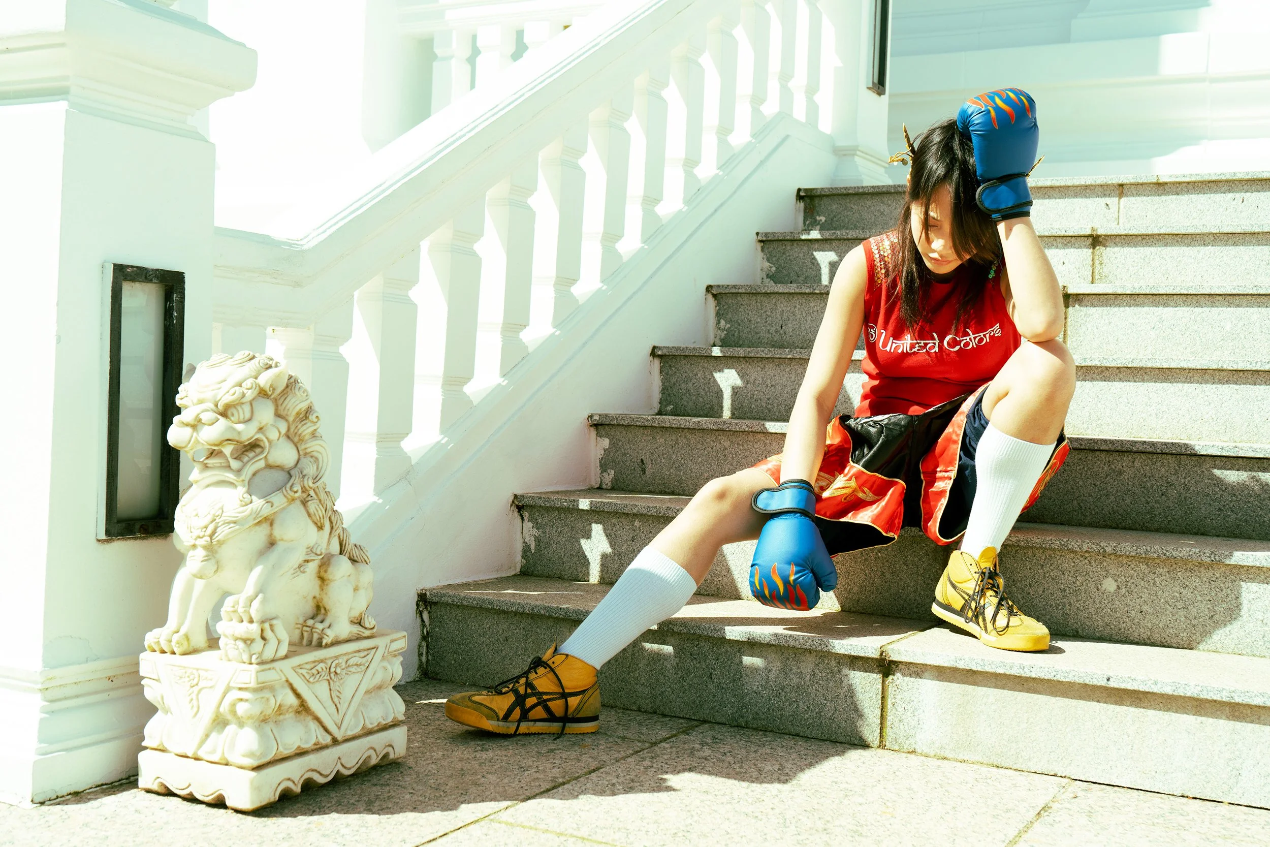 A girl dressed in colorful sportswear, including a red tank top, shorts, white knee-high socks, and yellow sneakers, is sitting on the stairs outside a building with white railing and a decorative stone sculpture nearby. She is wearing boxing gloves and appears to be tired or resting.