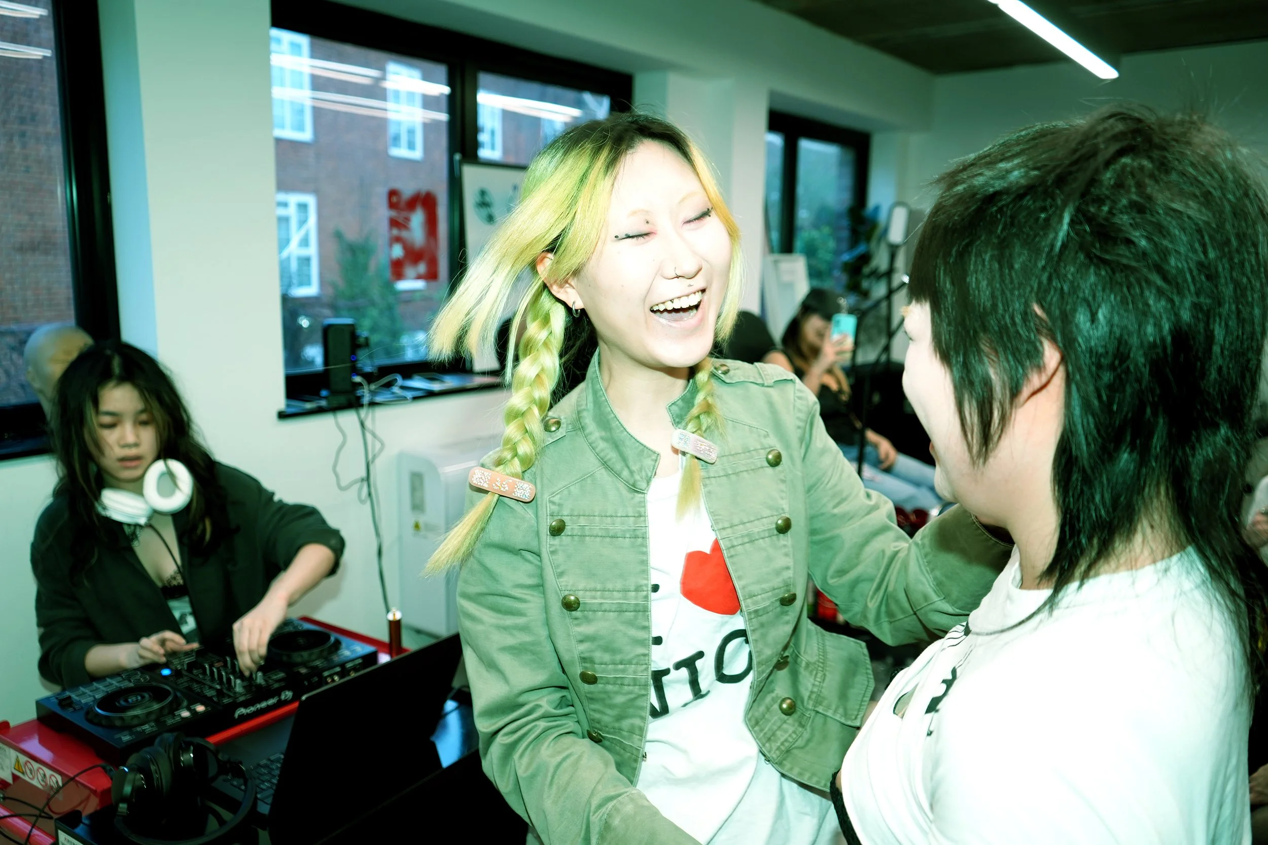 A young woman with blonde braided hair and a nose ring laughing and talking to a person with short black hair in a room with large windows, with people in the background including a girl with headphones and DJ equipment.